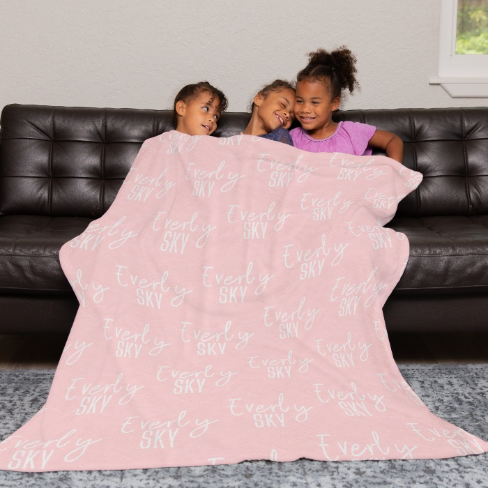 Three cheerful children huddle under a personalized pink baby blanket with 'Everly SKY' script, draped over a homey leather couch.