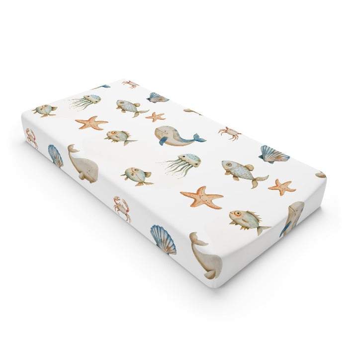 Eco-friendly changing pad cover with a marine-inspired design, showcasing vibrant illustrations of ocean creatures such as whales, fish, crabs, starfish, and jellyfish on a clean white base, perfect for environmentally conscious families.