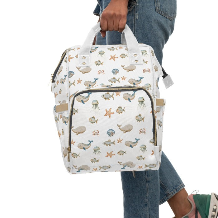 Bright and airy diaper backpack, with a white base highlighting a cheerful ocean motif including whales, fish, crabs, starfish, and jellyfish, perfect for parents who love the ocean