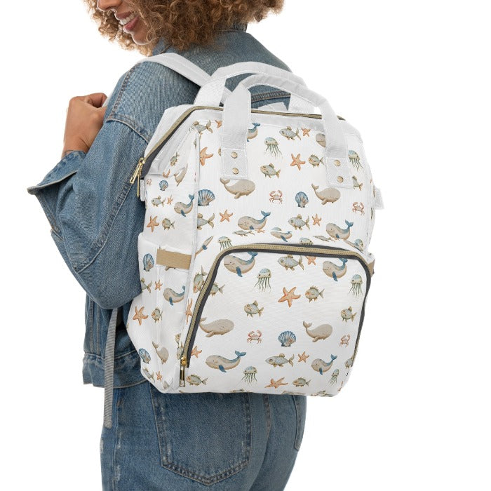 White-background diaper backpack with vibrant ocean-themed illustrations, including whales, fish, crabs, starfish, and jellyfish, combining functionality with a love for the sea.